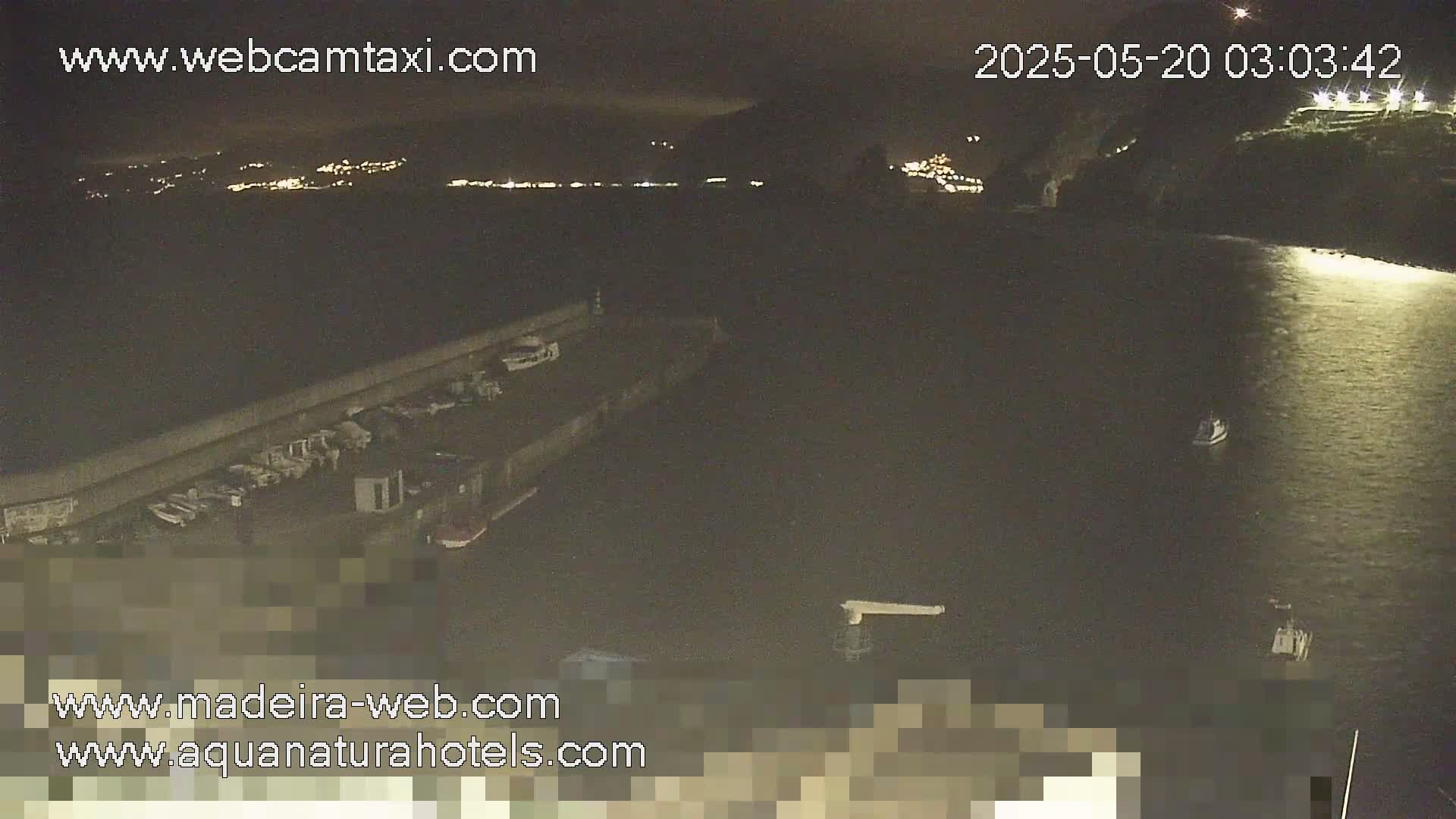A nighttime, low-light view of a harbor with boats moored alongside a long wall, and a few small boats on the dark water.