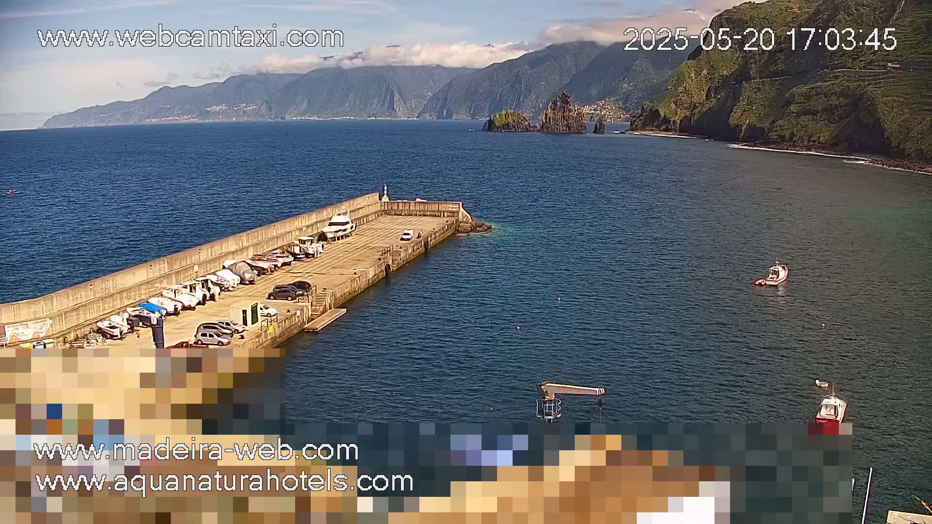 A small harbor with boats docked, a concrete wall, and calm blue water under a partly cloudy sky, with mountainous cliffs visible in the distance.