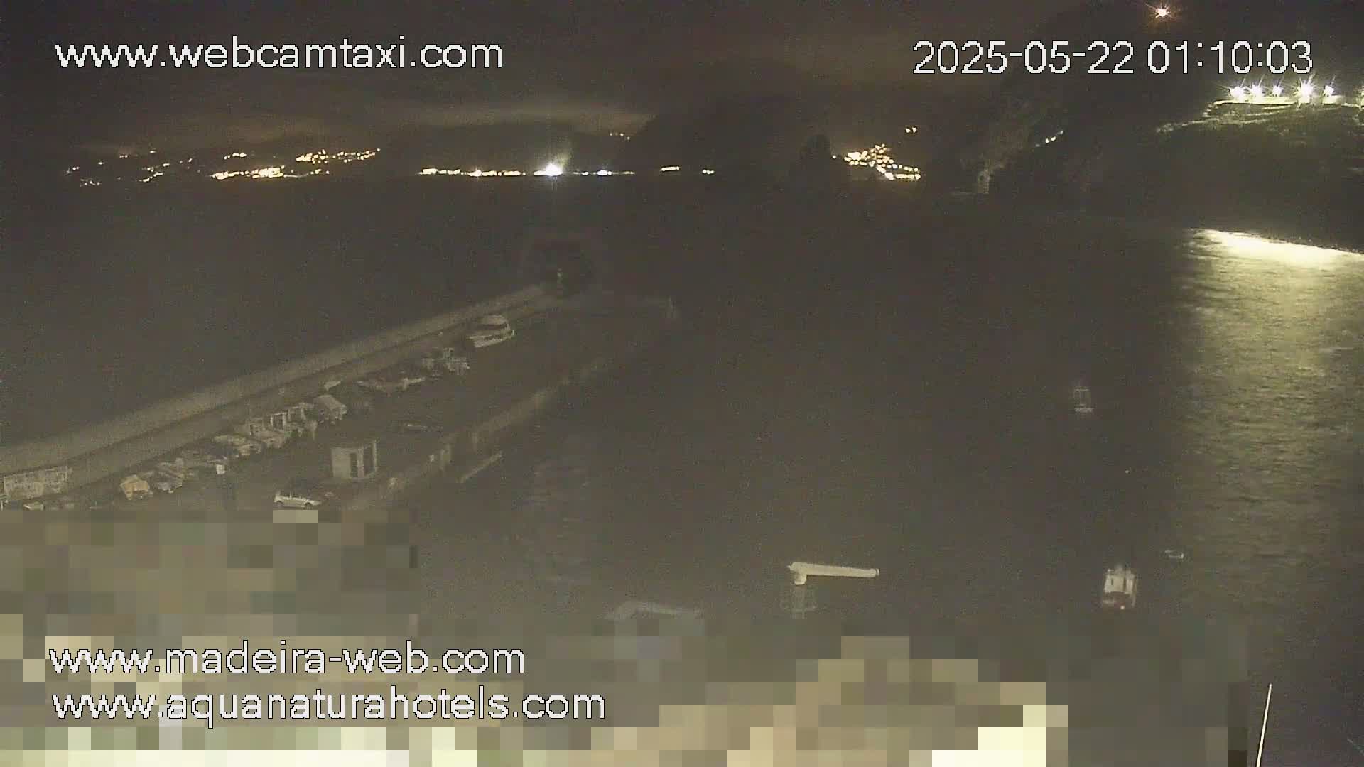 A nighttime view of a dark body of water next to a parking lot and a dimly lit shoreline, with some distant lights visible across the water.
