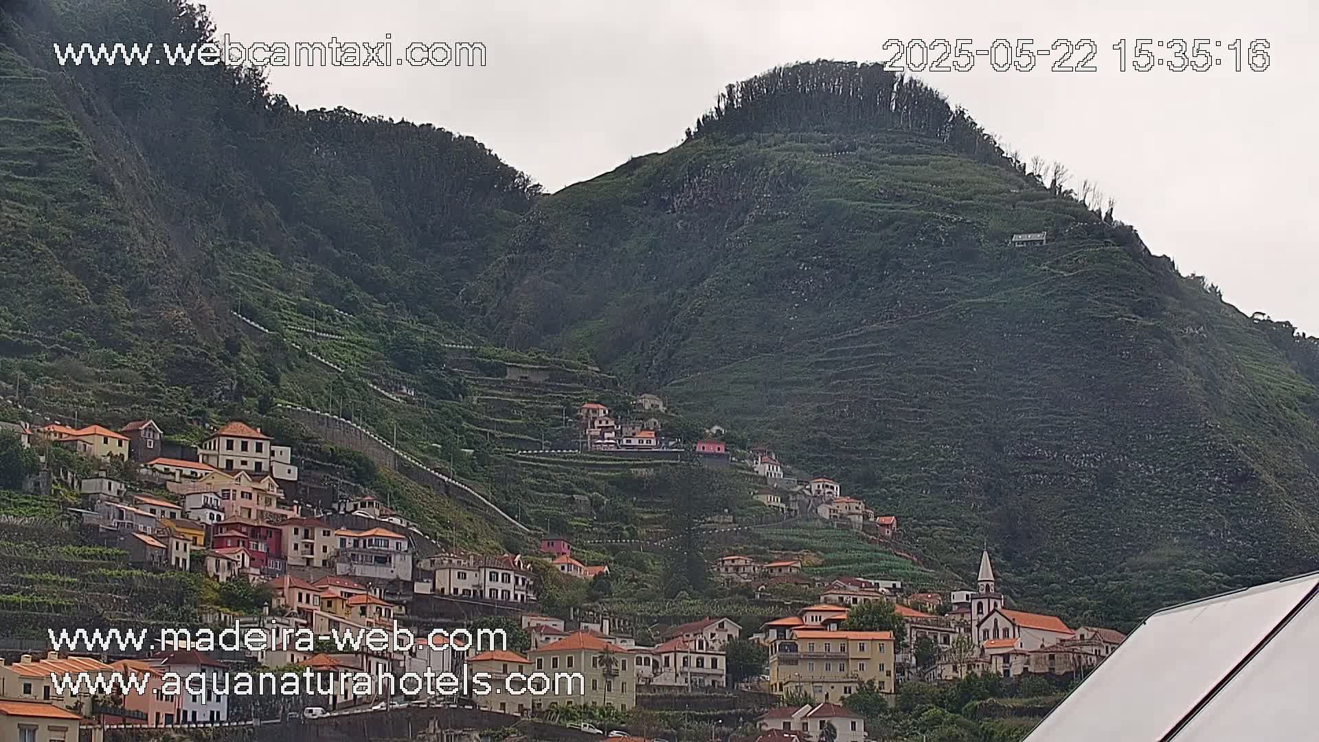A village of red-roofed houses clings to the side of a verdant, terraced hillside under an overcast sky.