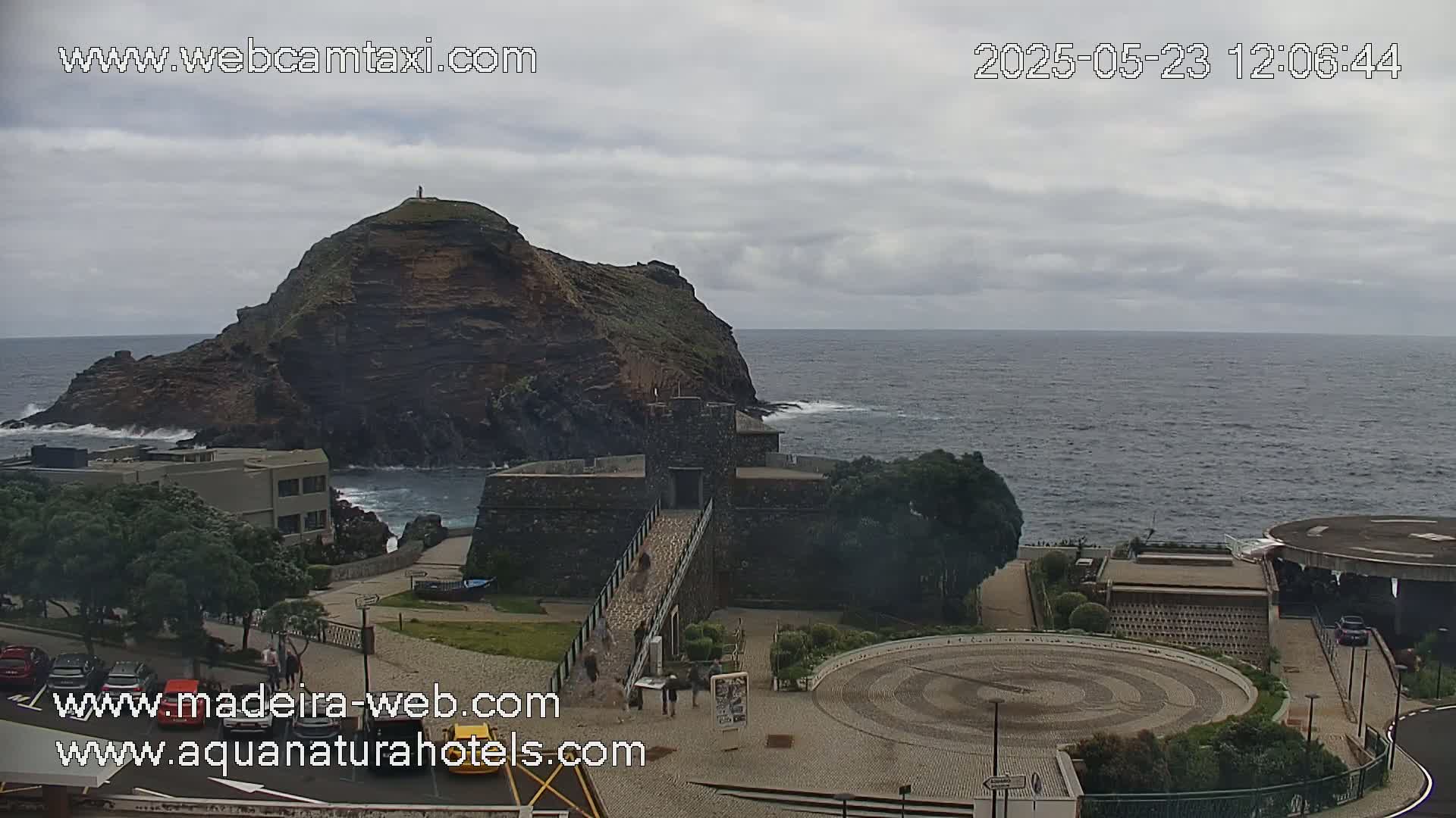 An overcast day reveals a coastal scene with a rocky islet, a stone structure resembling a fortress, and a paved area with a circular design, all overlooking a dark-blue ocean with some waves.
