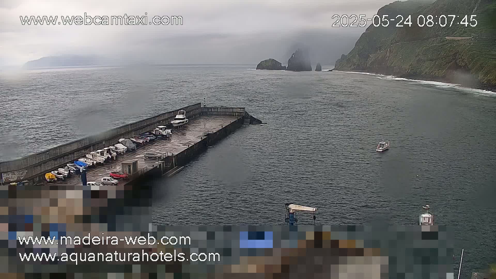 A foggy day shows a harbor with several boats docked and a few more in the water, near a rocky cliffside.