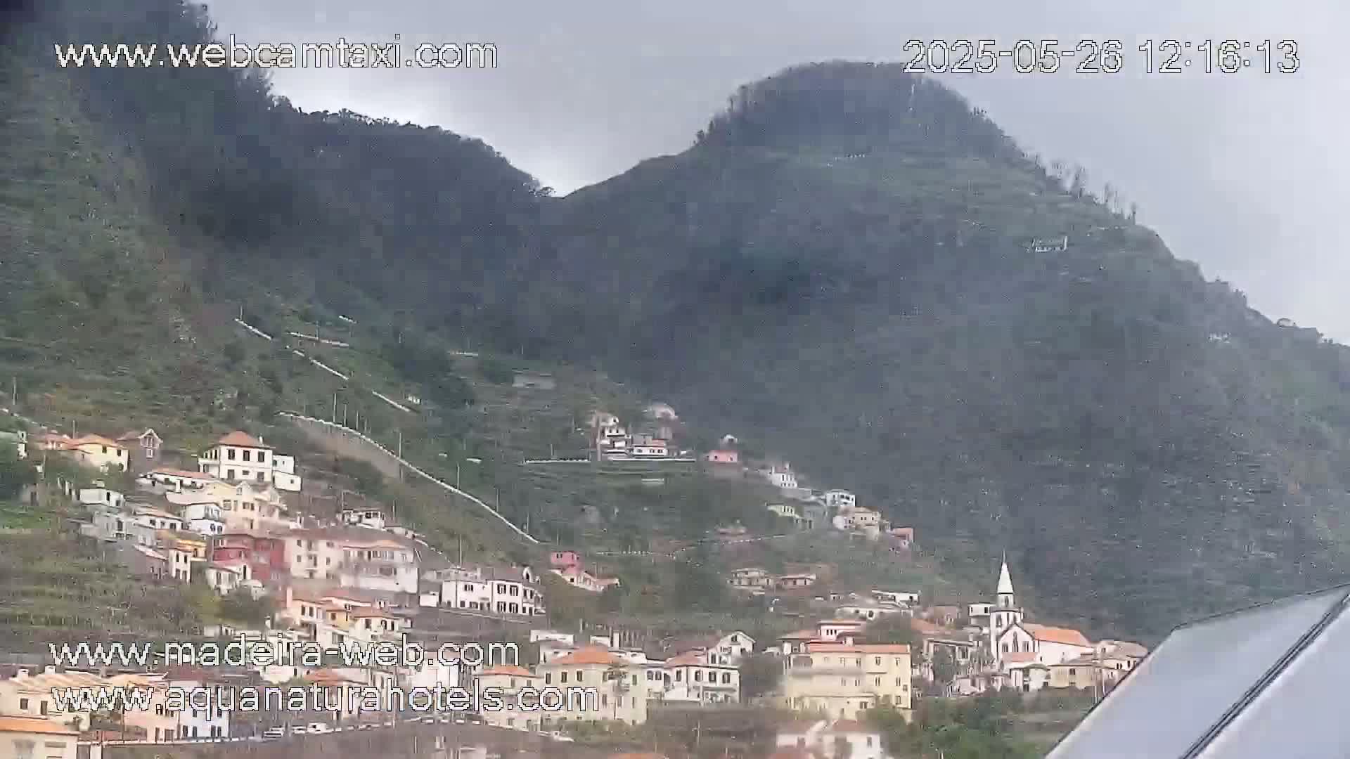 A village clings to the slopes of two steep, dark green hills under an overcast sky.