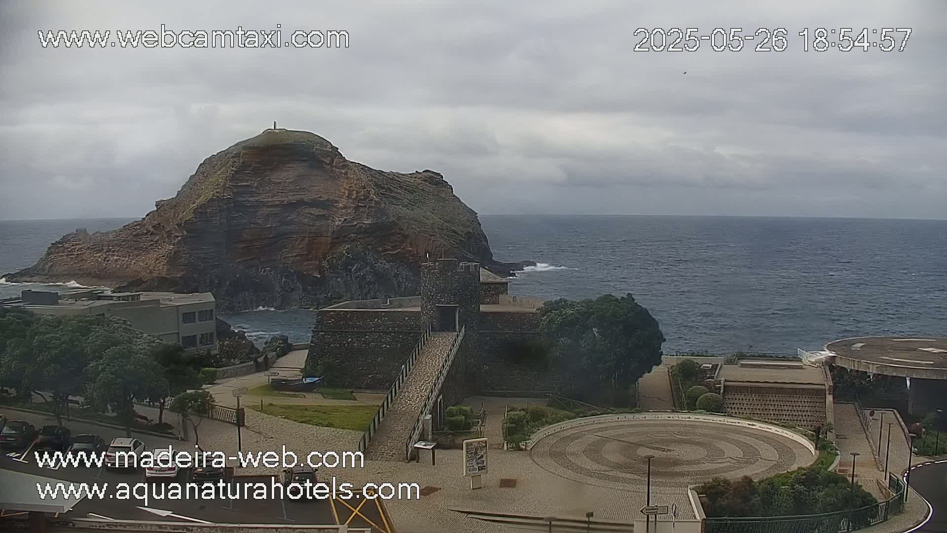A rocky cliff overlooks a calm ocean under a cloudy sky, with a stone structure and paved areas situated below.