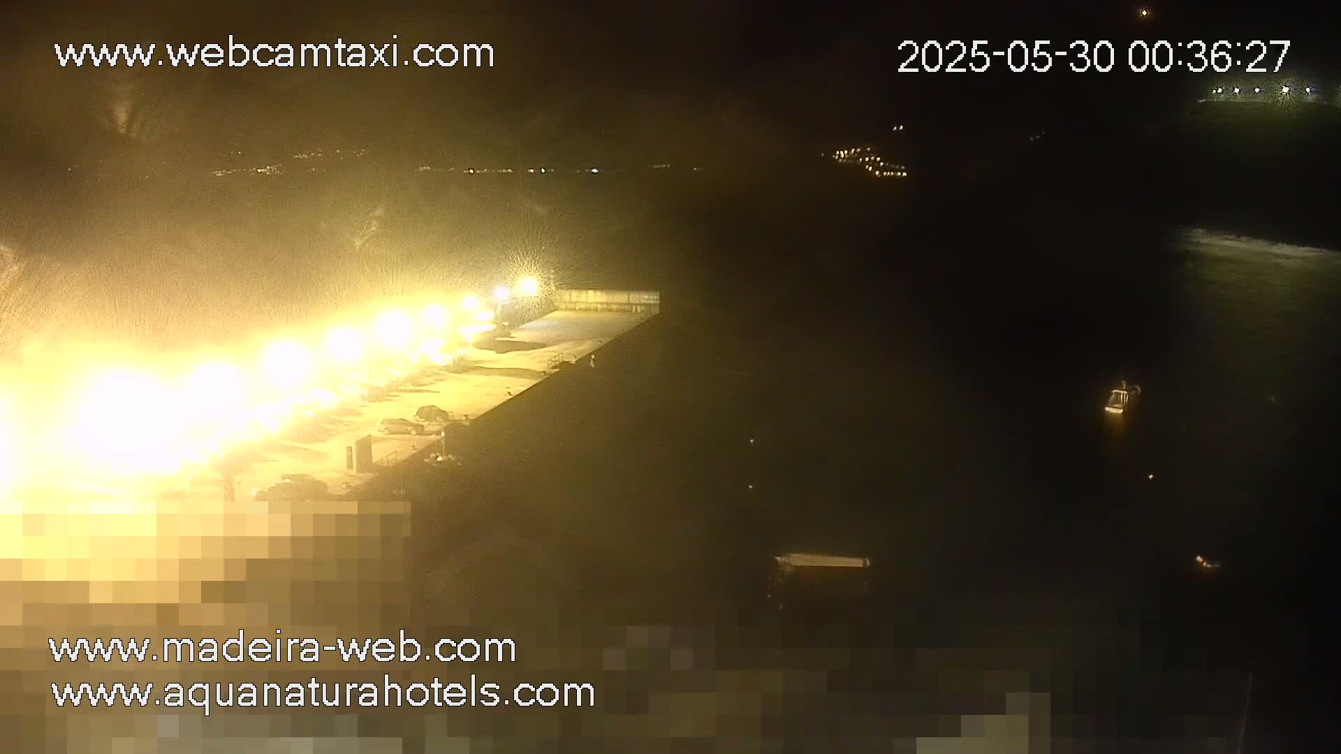 A nighttime, brightly lit waterfront area with a body of dark water and a few small vessels visible in the distance.