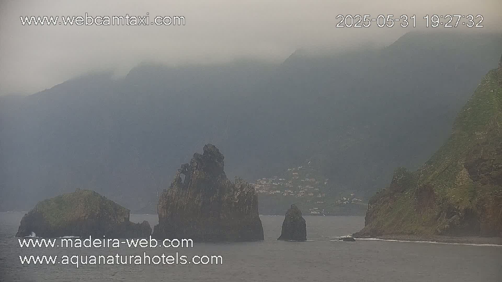 A foggy coastal scene shows dark, rocky formations in the sea near a hazy green hillside and a small village.