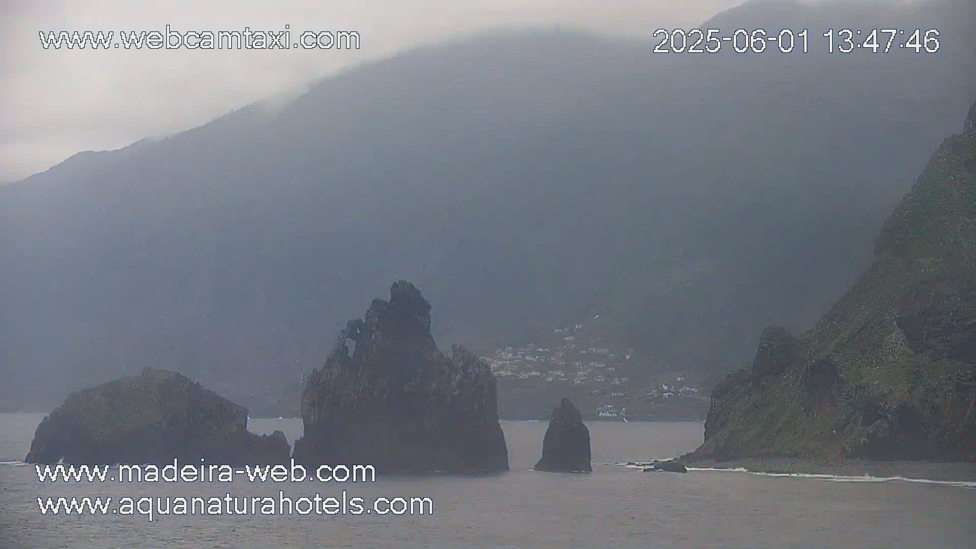 A hazy, overcast day reveals dark, rocky outcrops in the ocean near a distant, partially visible village nestled between steep, green hillsides.