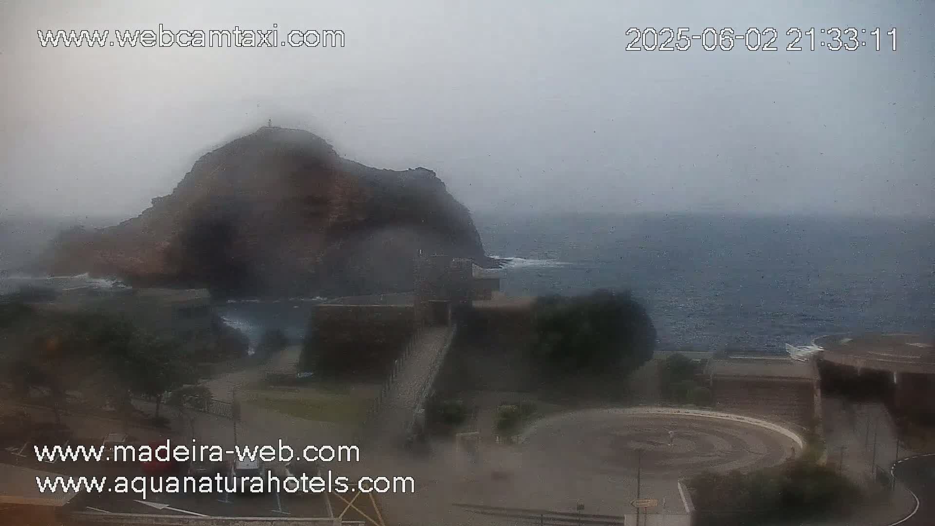 A hazy, overcast day shows a rocky cliff overlooking a dark, wavy ocean, with a paved area and some buildings in the foreground.