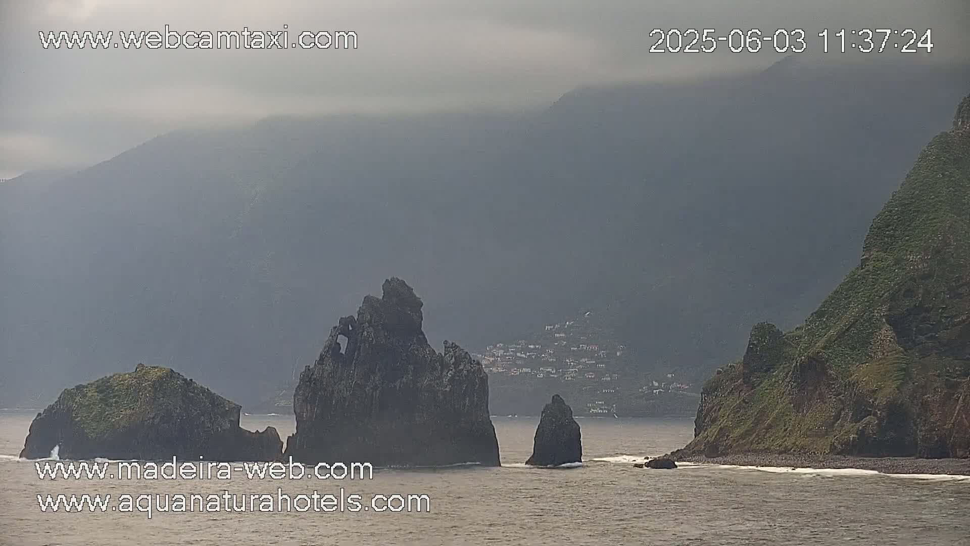 A hazy, overcast day reveals several dark, rocky sea stacks in the ocean near a mountainous coastline.