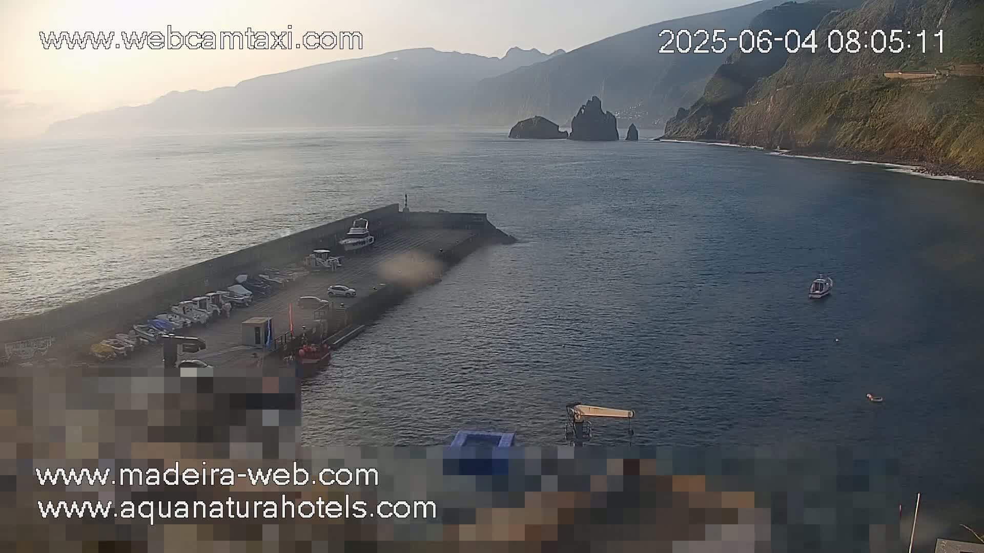 A hazy sunrise illuminates a harbor with several boats docked, a coastline with dark, rocky cliffs, and calm waters.