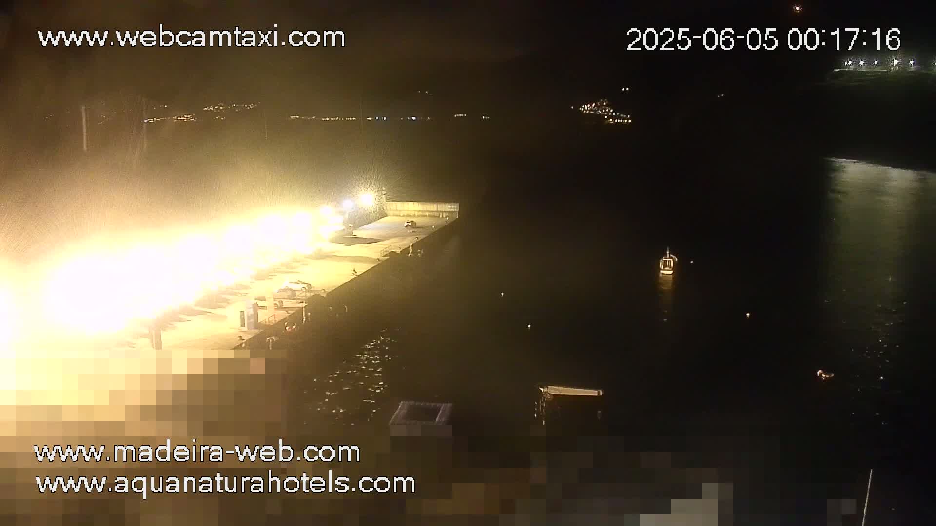A brightly lit dock at night is seen from above, next to a dark body of water with a few small boats.
