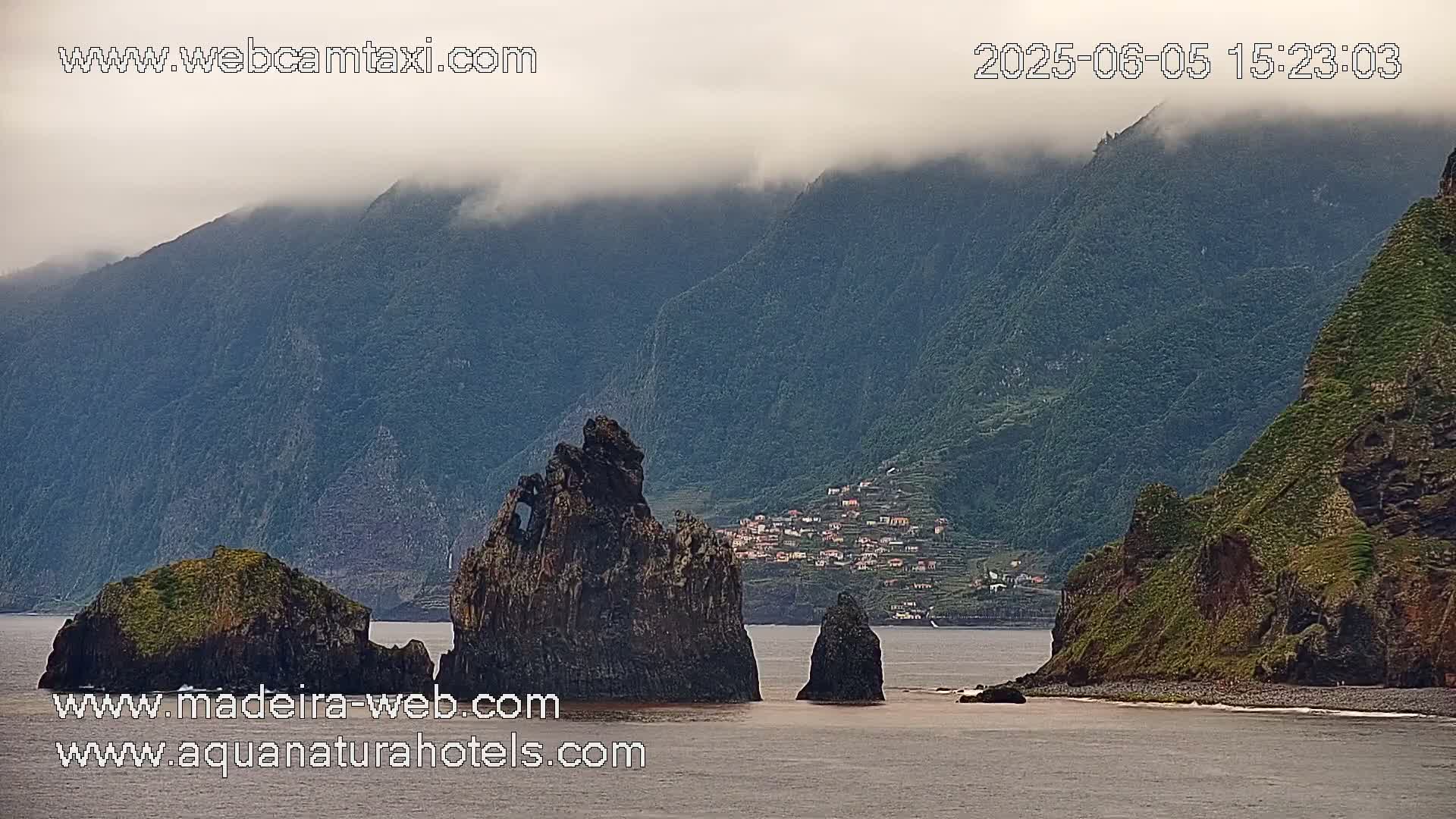 A coastal scene shows dark, rocky formations in the foreground, with a village nestled in the hillside of a large, foggy mountain in the background.