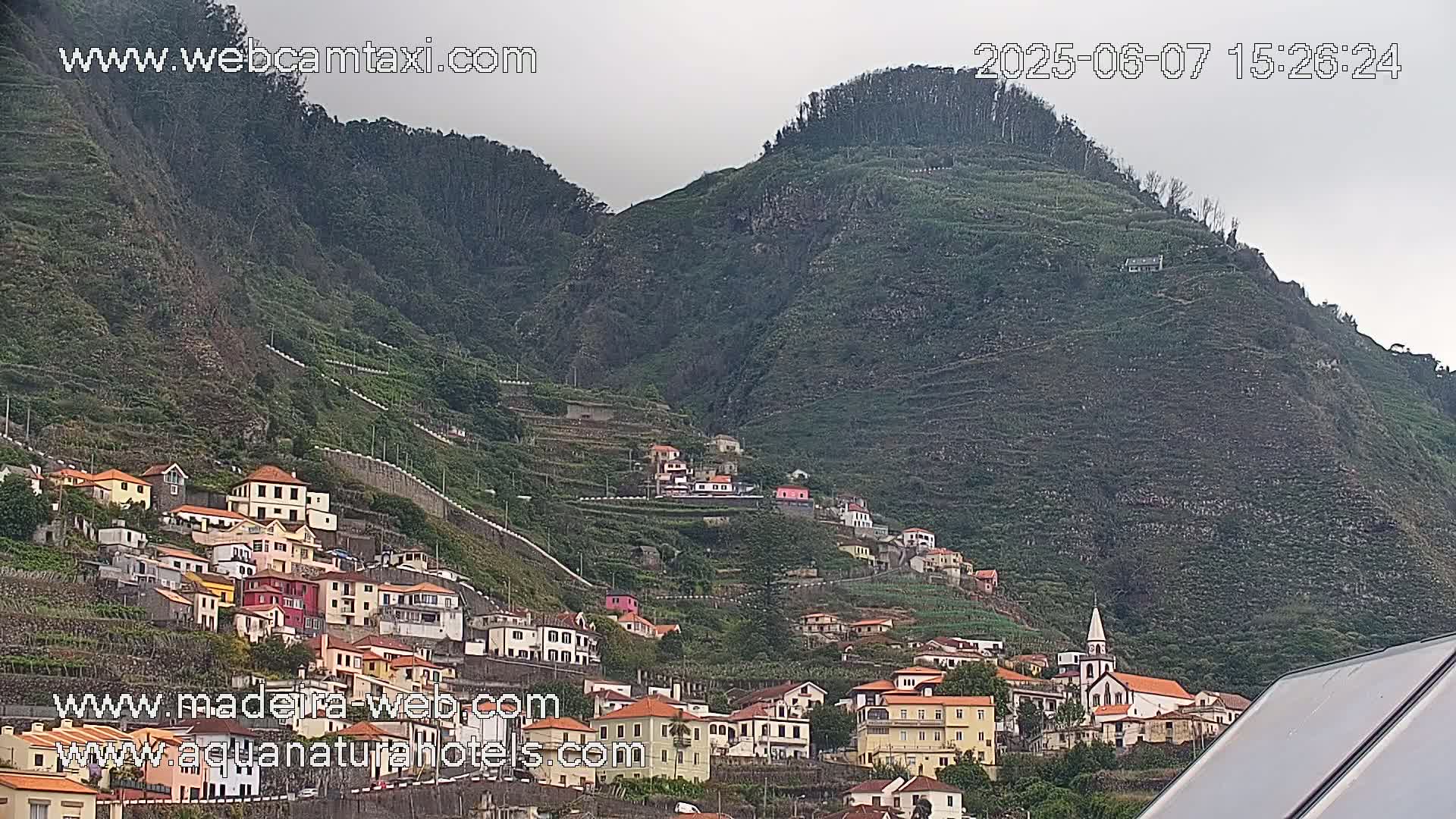 A village of colorful houses climbs a steep, verdant hillside under an overcast sky.