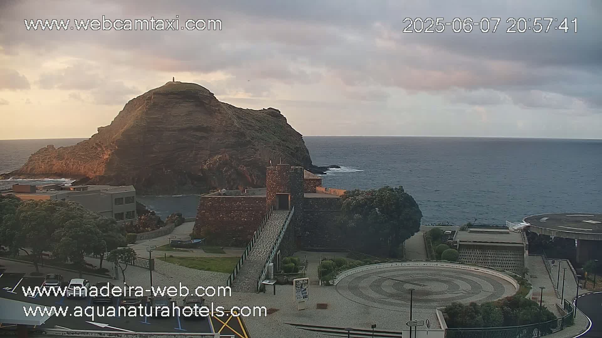 A rocky headland juts into a calm ocean under a partly cloudy sky, with a stone structure and paved plaza in the foreground.