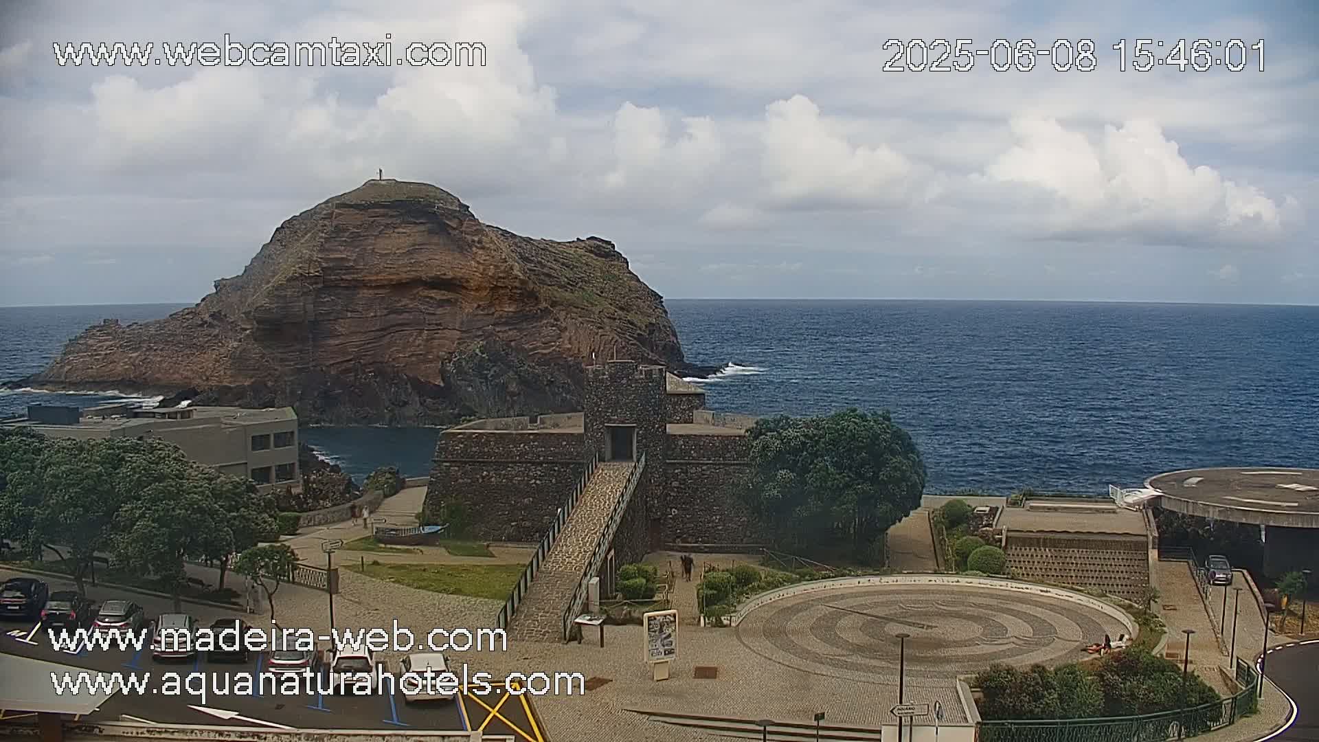 A rocky island is visible from a coastal viewpoint, under partly cloudy skies, with a stone structure and paved plaza in the foreground.