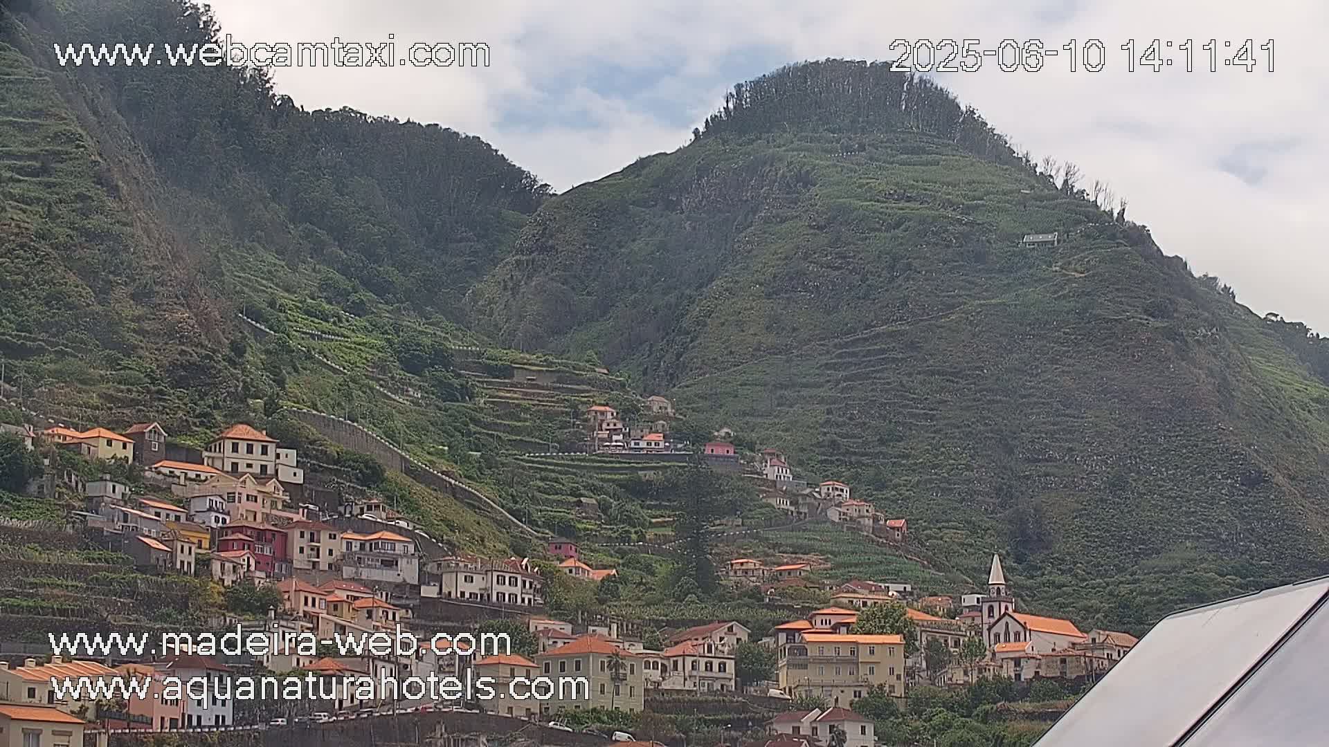 A village of low, light-colored buildings clings to the side of a steep, terraced, green mountain under a partly cloudy sky.