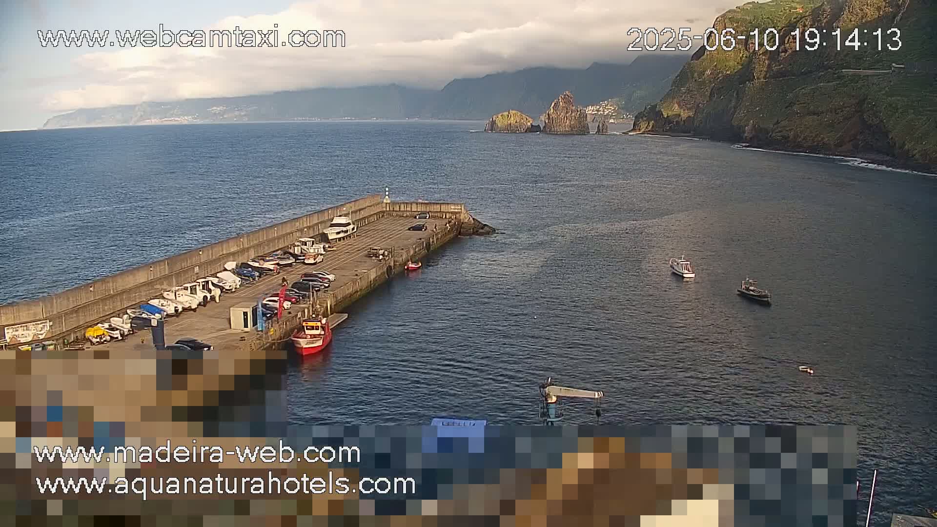 A harbor with several small boats and cars parked along a concrete wall, situated near a rocky coastline under partly cloudy skies.