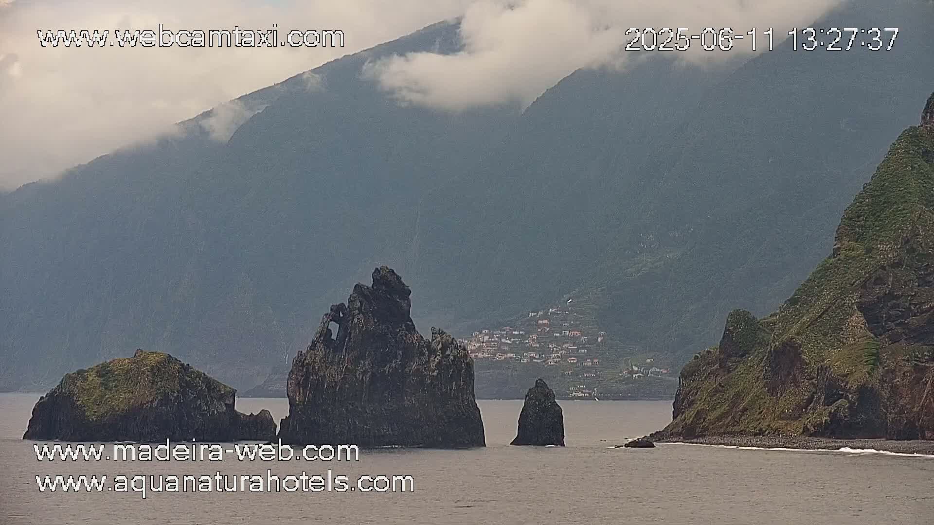 A cloudy coastal scene shows dark, rocky outcroppings in the ocean near a mountainous shoreline with a small village nestled on the hillside.