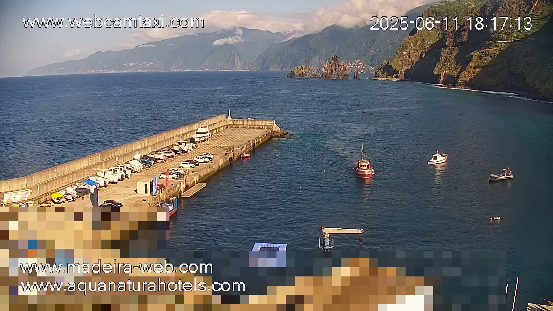 A small harbor with boats docked and parked cars, situated next to a calm ocean with mountainous cliffs in the background under a partly cloudy sky.