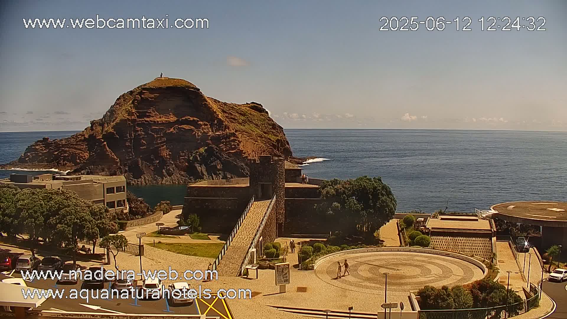 A coastal scene on a sunny day shows a rocky, reddish-brown hill overlooking a calm ocean, with a stone structure and paved plaza in the foreground.