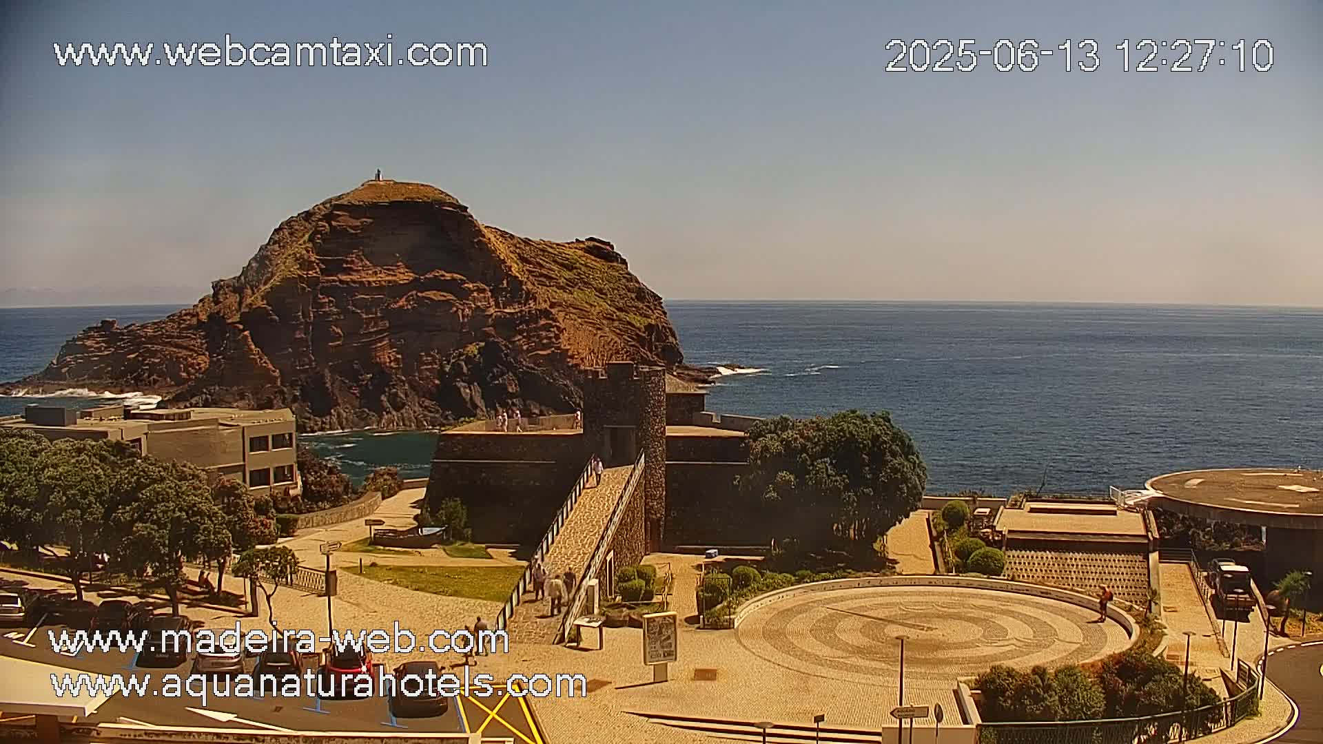 A rocky, reddish-brown island juts out into a calm, blue ocean under a clear, sunny sky, with a stone structure and paved plaza visible on the shore.