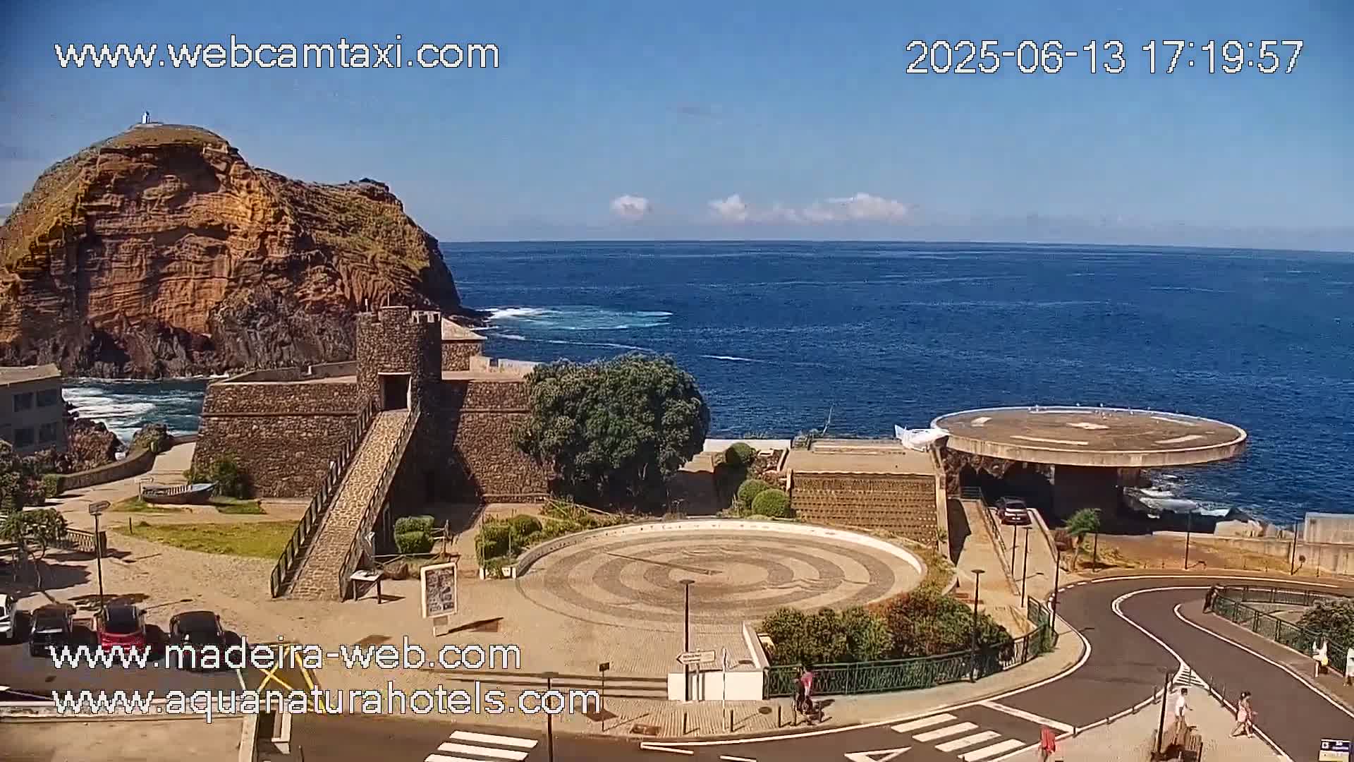 A coastal scene with a stone fortress, a circular plaza, and a helipad overlooks a calm ocean under a mostly sunny sky.