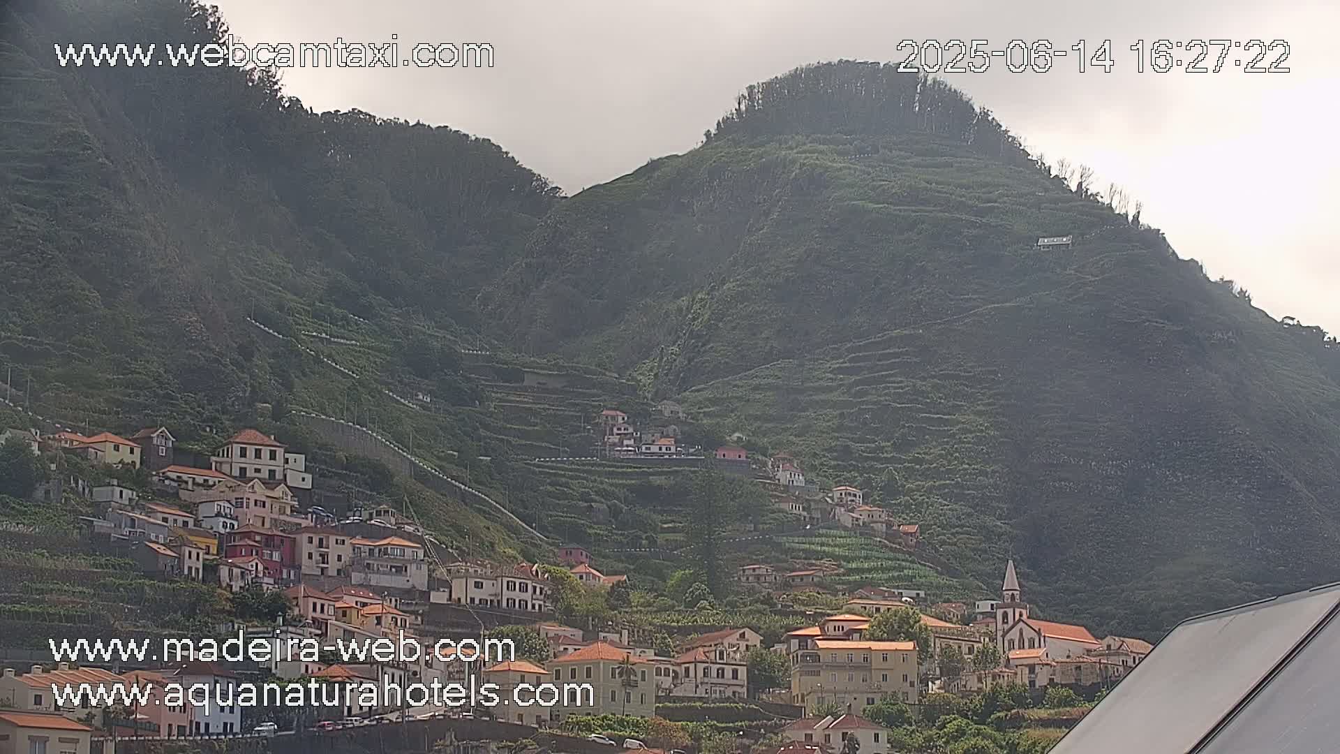A hillside village clings to the slopes of a verdant, terraced mountain under an overcast sky.