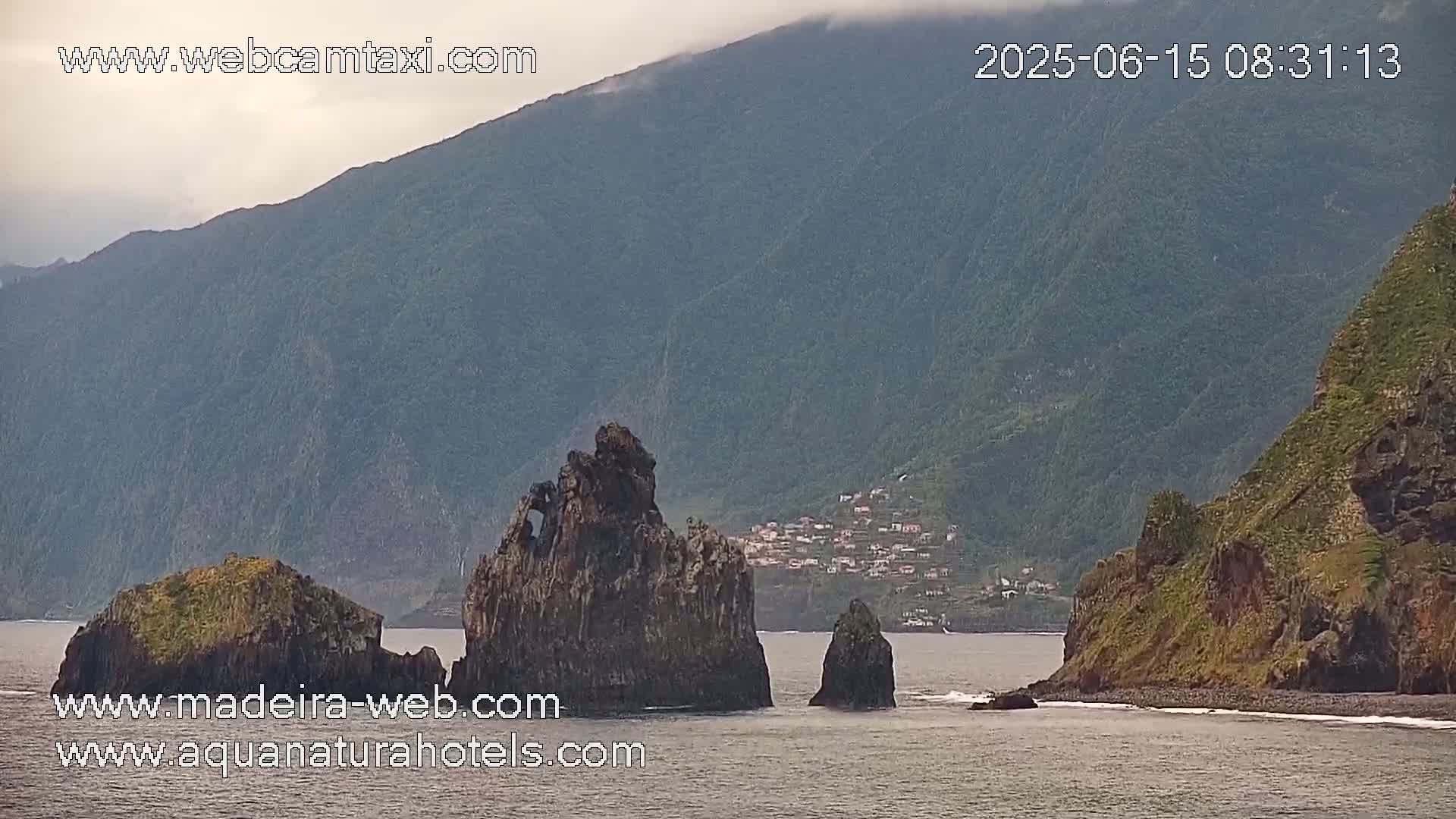 Ocean waves lap against a rocky shore, where several dark, tall rock formations rise from the water near a small village nestled in the foothills of a large, misty mountain.