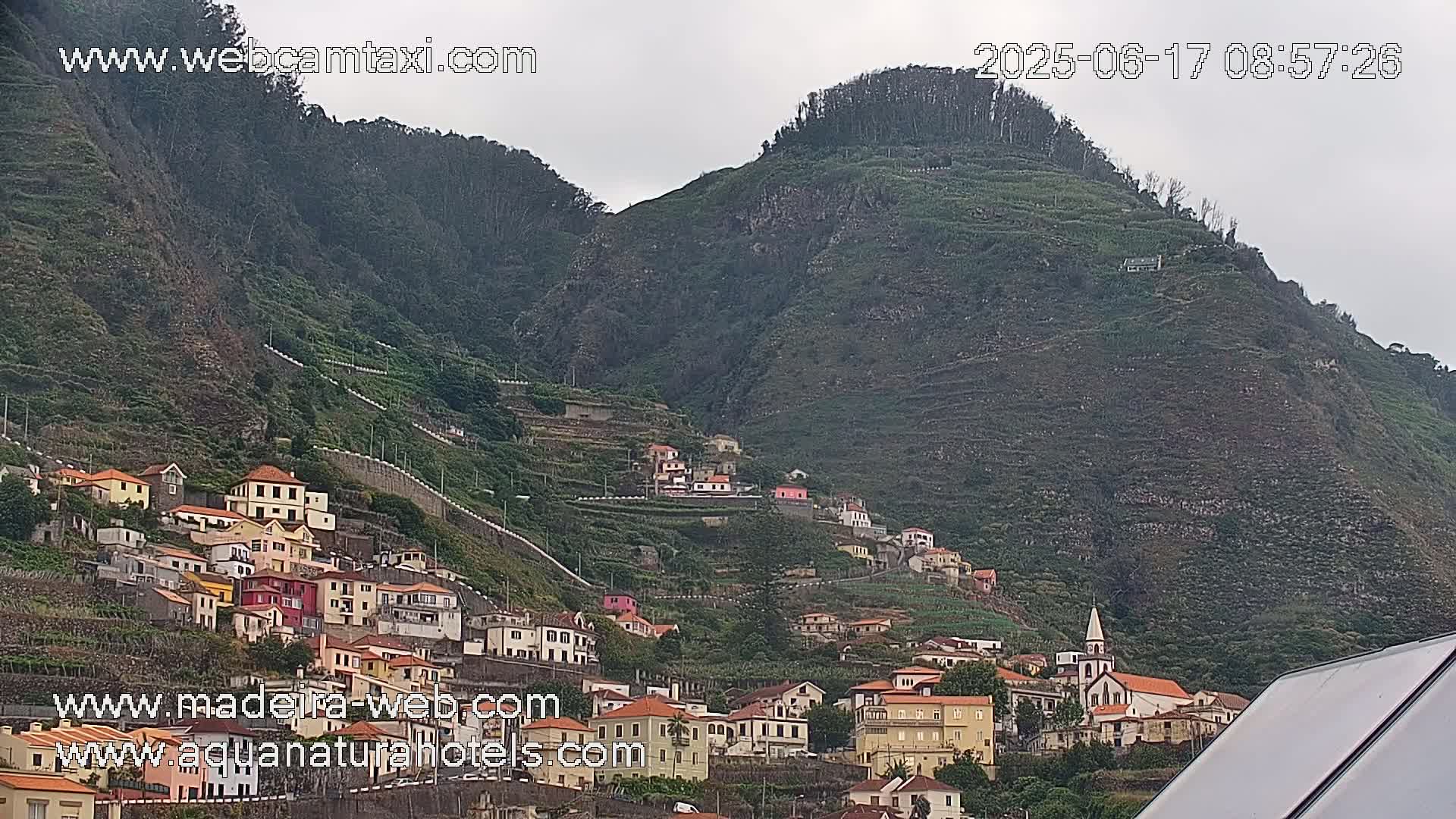 A village of colorful buildings clings to the steep, green slopes of a mountain under an overcast sky.