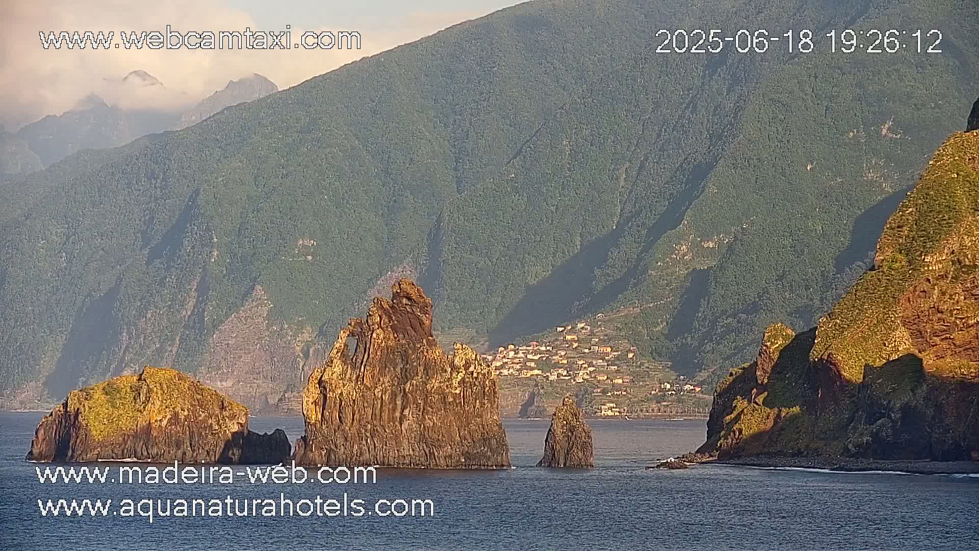 A coastal scene shows several rocky outcroppings in calm water near a village nestled in a green, mountainous landscape under a mostly clear sky.