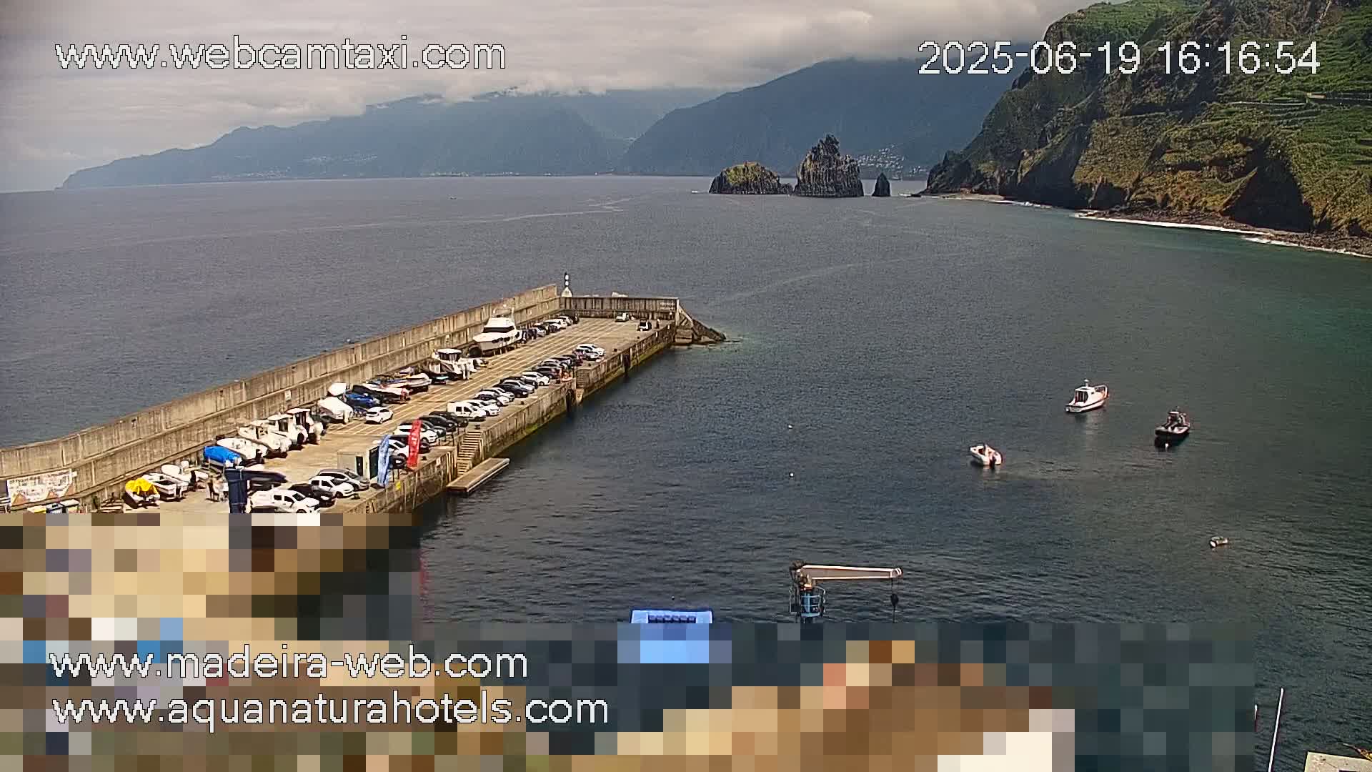 A harbor with parked cars and boats, under partly cloudy skies, overlooks a calm ocean with distant mountains and a few small boats in the water.