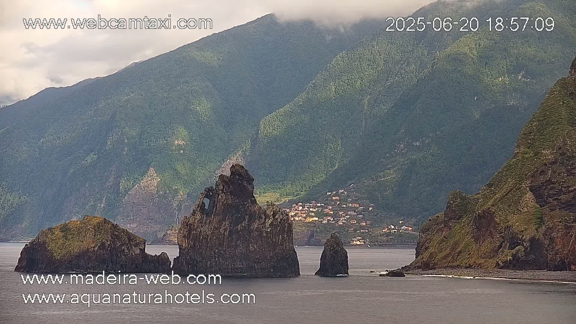 A mostly cloudy day shows dark, rocky, outcroppings in the ocean near a village nestled in green hills.
