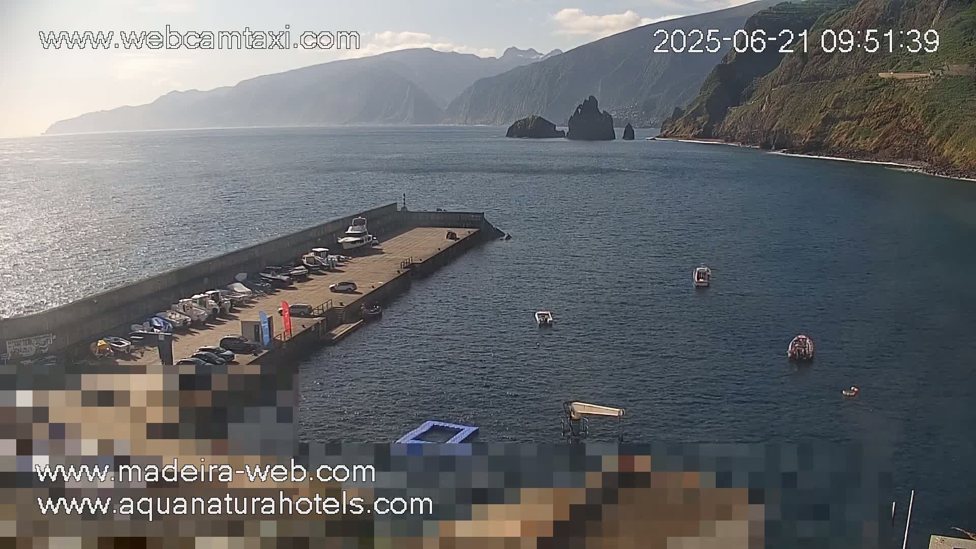 A harbor with several boats docked and in the water, next to a rocky coastline under sunny skies.