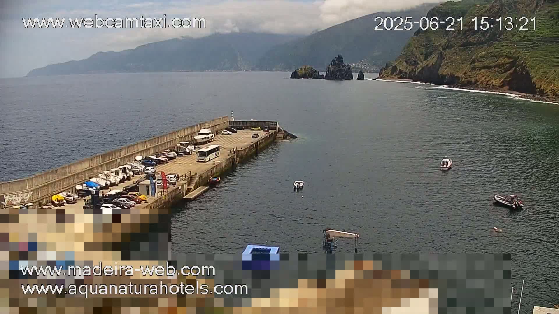 A harbor with boats and cars parked along a concrete pier, under partly cloudy skies, and a rocky coastline in the background.