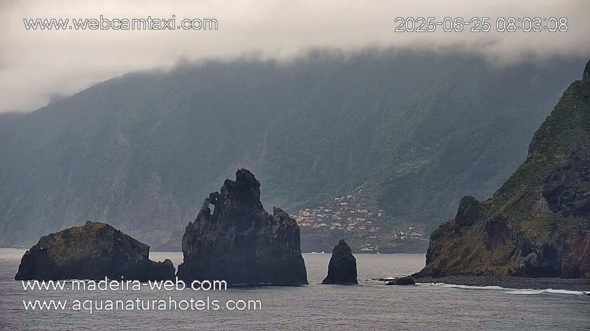 A foggy coastal scene shows dark, rocky cliffs and islands, with a small village visible on a hillside in the background.