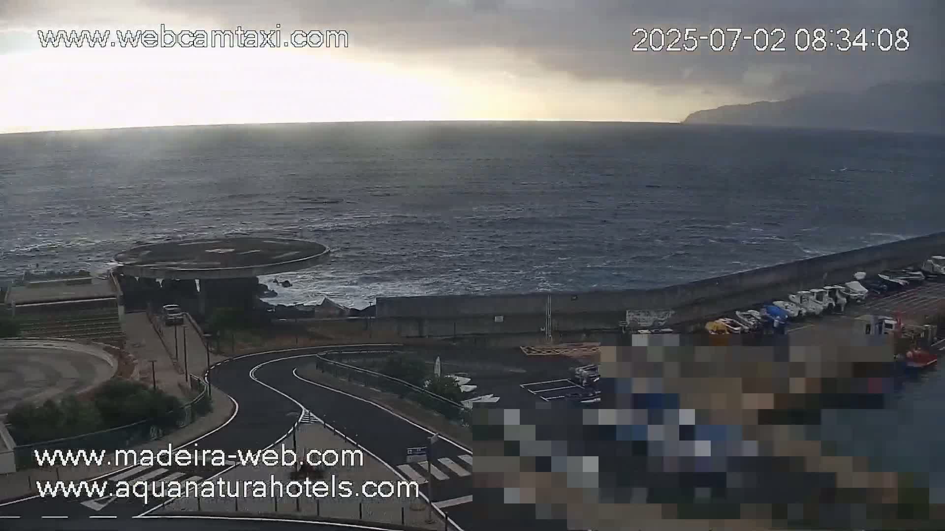 A mostly cloudy day shows a choppy ocean meeting a seawall with parked boats and a winding road leading to a circular structure.