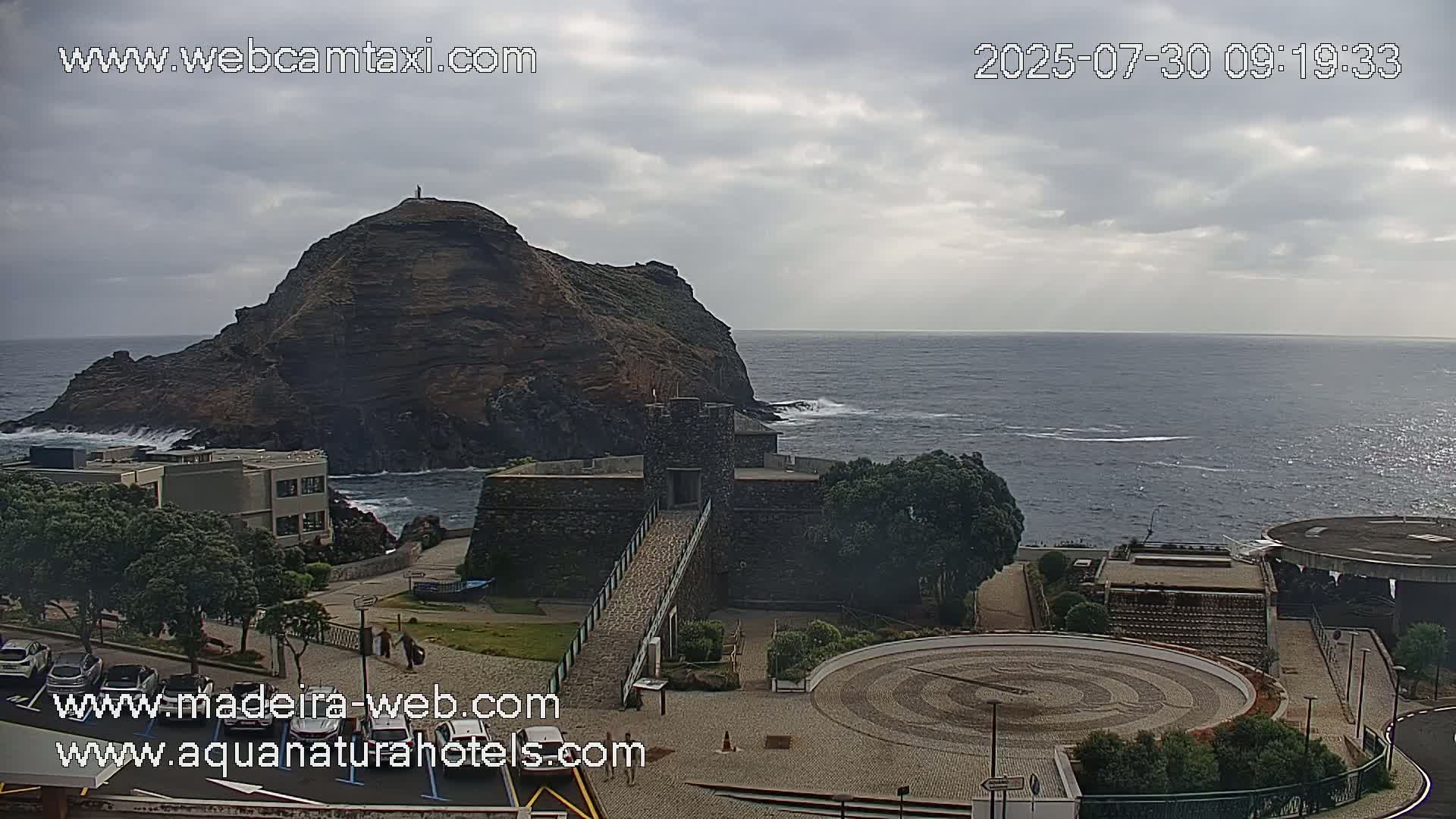 A coastal scene under a cloudy sky shows a rocky hill, a stone structure with a walkway, a paved plaza with a circular design, and a body of water with some wave action.