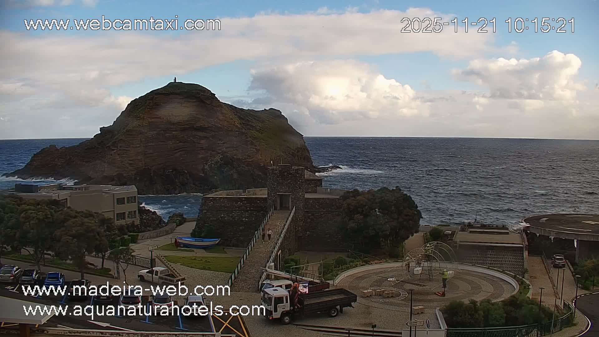 The image displays a rugged coastal landscape featuring a prominent rocky headland overlooking a turbulent ocean, with a stone fort, modern buildings, a helipad, and a circular paved plaza with a decorative structure in the foreground, all under a partly cloudy sky.