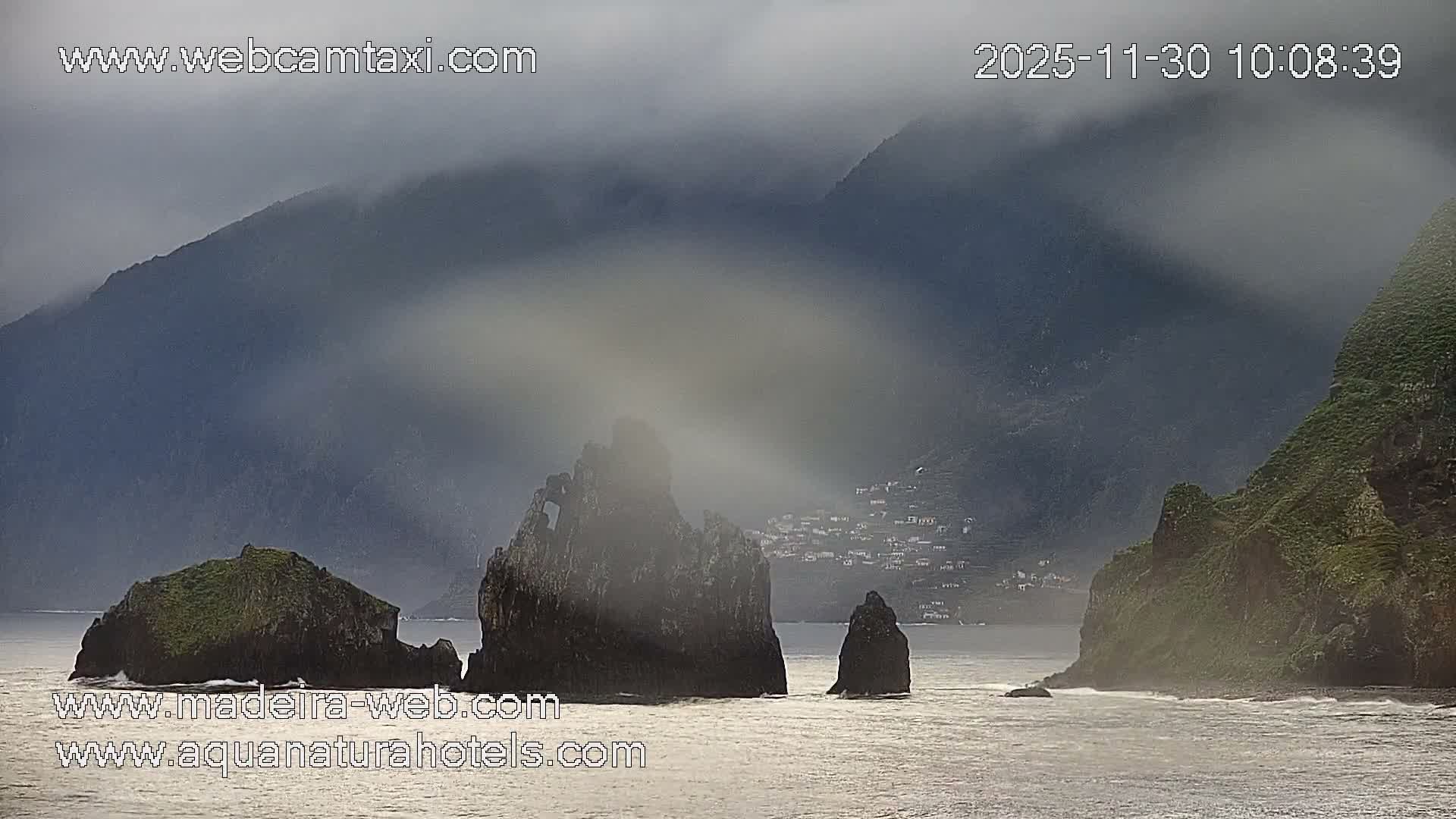 A rugged coastline is depicted on a misty, overcast day with several dark sea stacks emerging from the choppy ocean, steep green mountains partially obscured by clouds, and a small village visible along the distant shore.