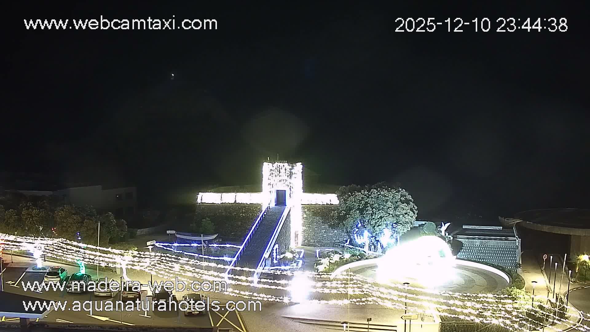 A clear night sky overlooks an outdoor scene brightly decorated with white lights, illuminating a cross-shaped building, a large fountain, and various trees.
