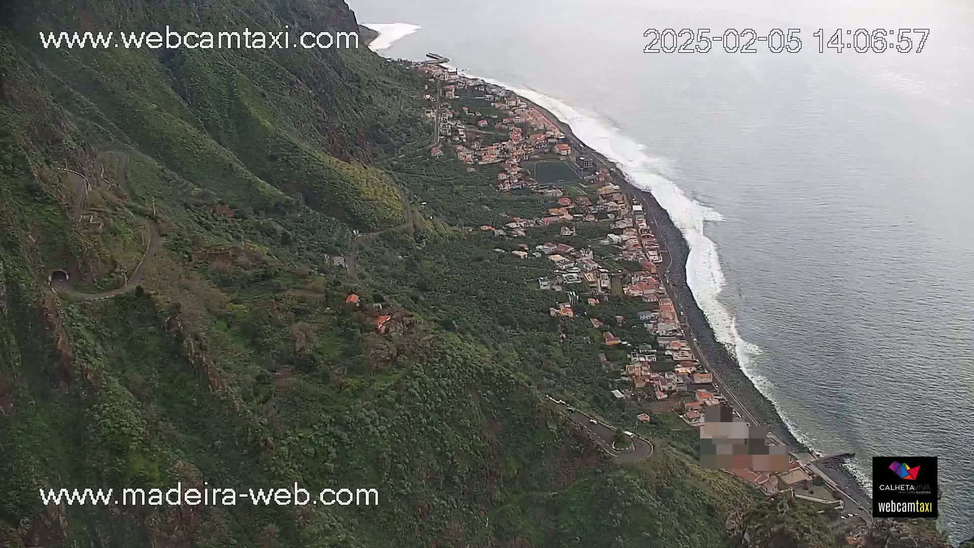 A coastal town nestled between a steep, green hillside and a dark-blue ocean with white-capped waves under an overcast sky.