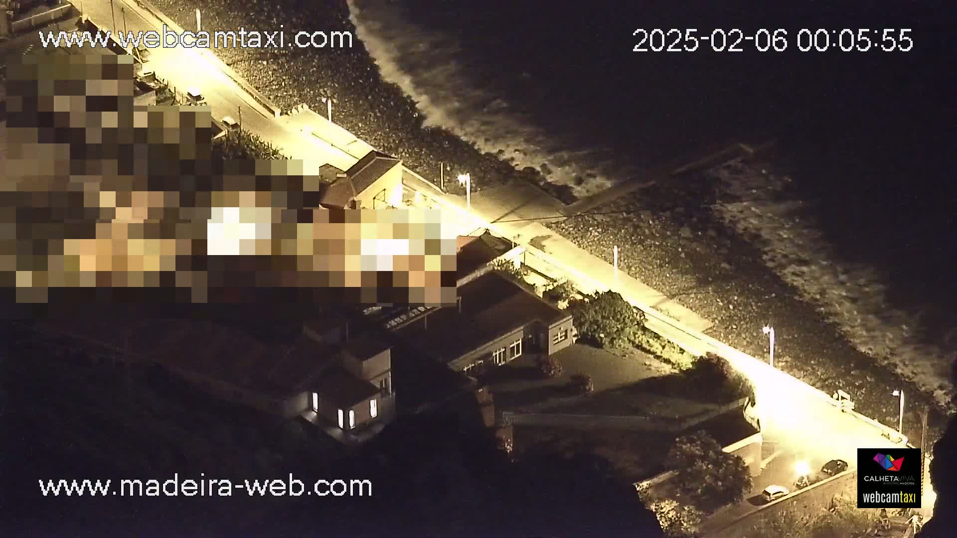 A nighttime aerial view shows a coastline with crashing waves, a brightly lit walkway, and several buildings along the shore.