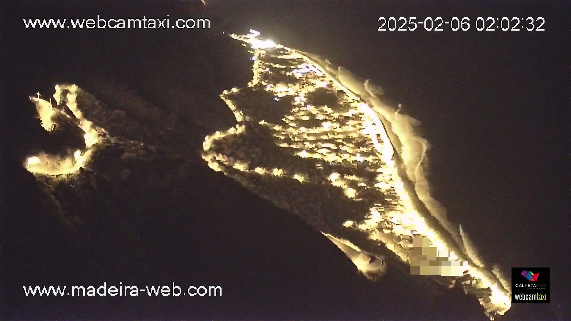 An aerial nighttime view shows a coastal town illuminated, with dark water and a sandy shoreline visible.