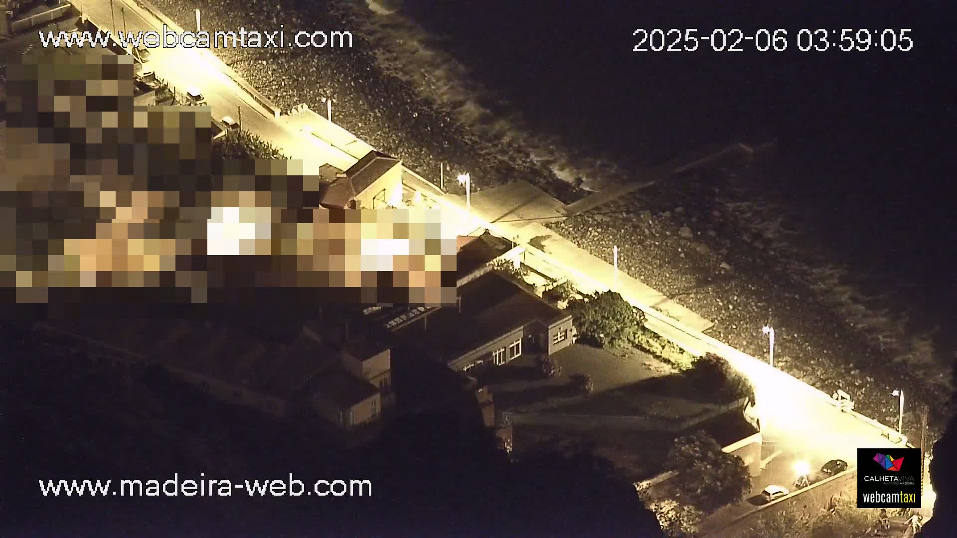 An aerial nighttime view shows a coastline with dark, wavy water, a lit paved walkway, and buildings along the shore.