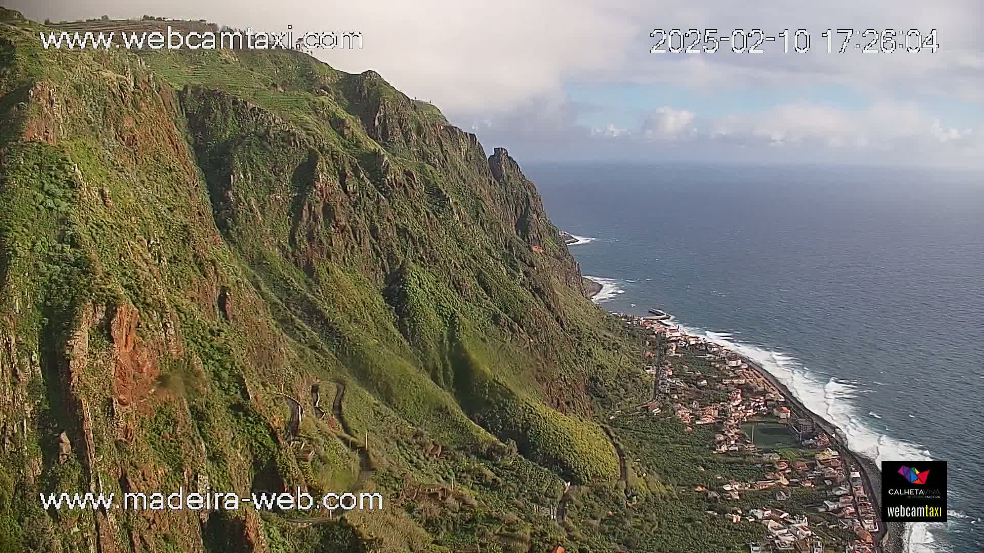A partly cloudy day reveals a steep, verdant cliffside overlooking a coastal town and the ocean, with waves breaking on the shore.