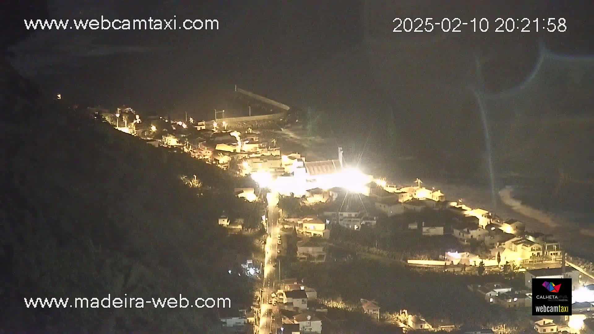 A nighttime, aerial view of a coastal town illuminated by streetlights, with dark, possibly cloudy, skies above.