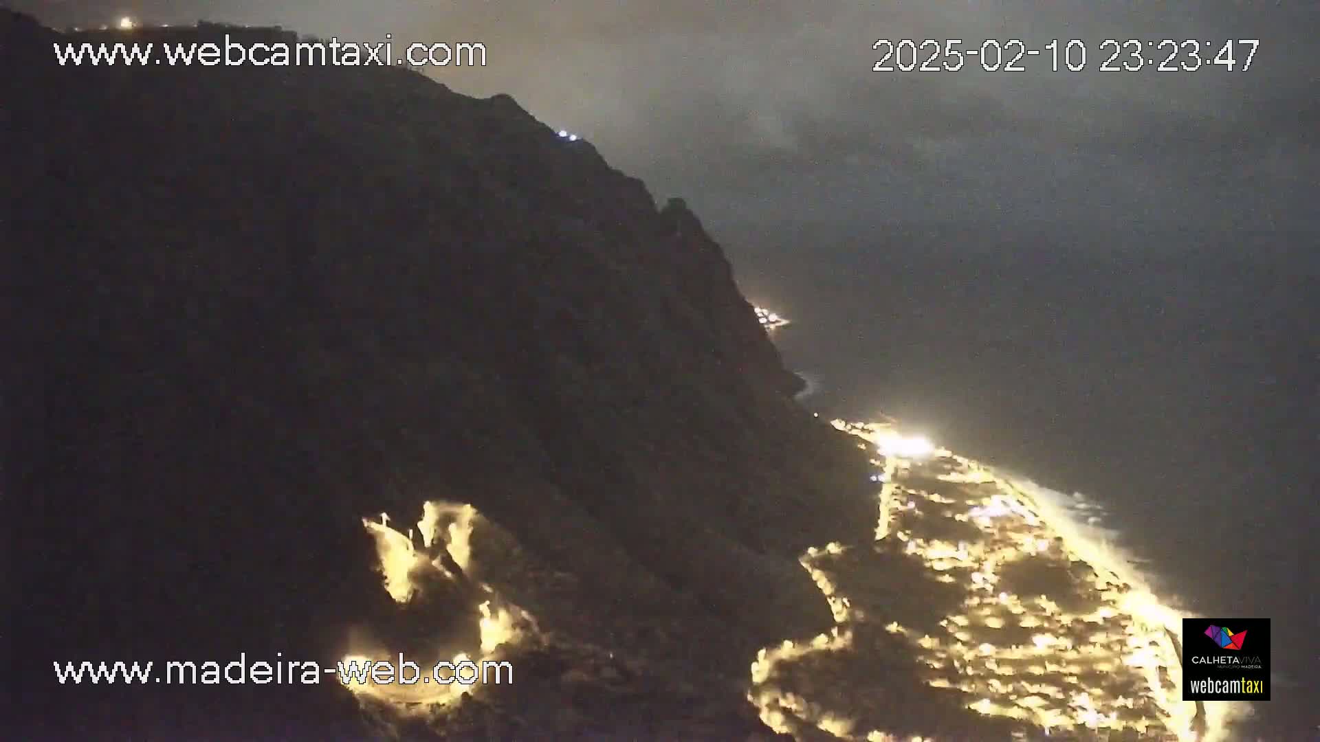 A nighttime view shows a dark cliffside overlooking a coastline illuminated by numerous lights.