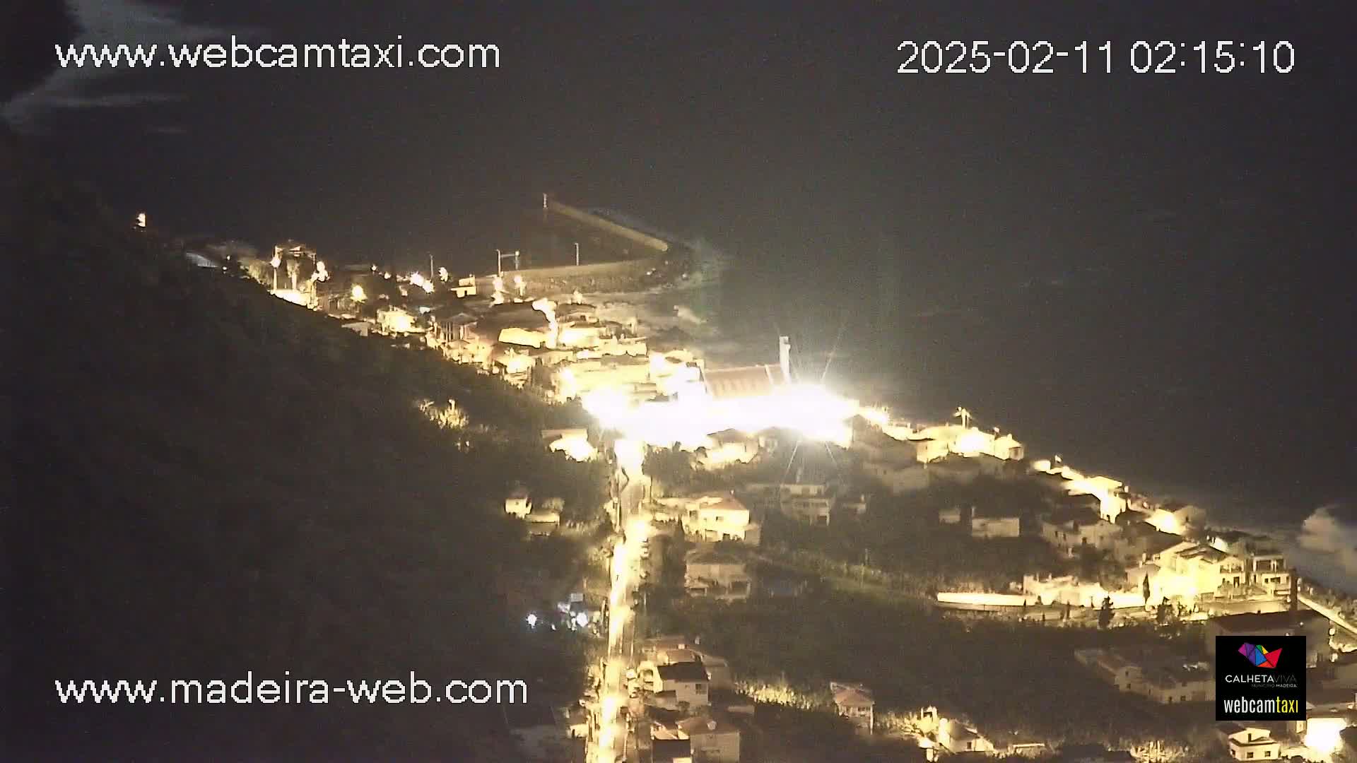 A nighttime, aerial view shows a coastal town illuminated by streetlights, with dark water visible below.