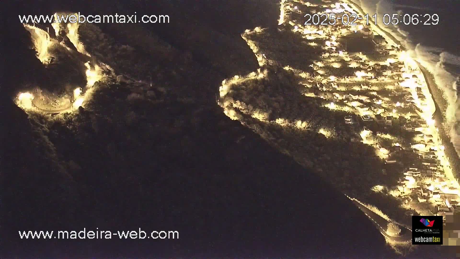 An aerial nighttime view shows a coastal town illuminated, with a winding road visible along the hillside and ocean waves breaking on the shore.