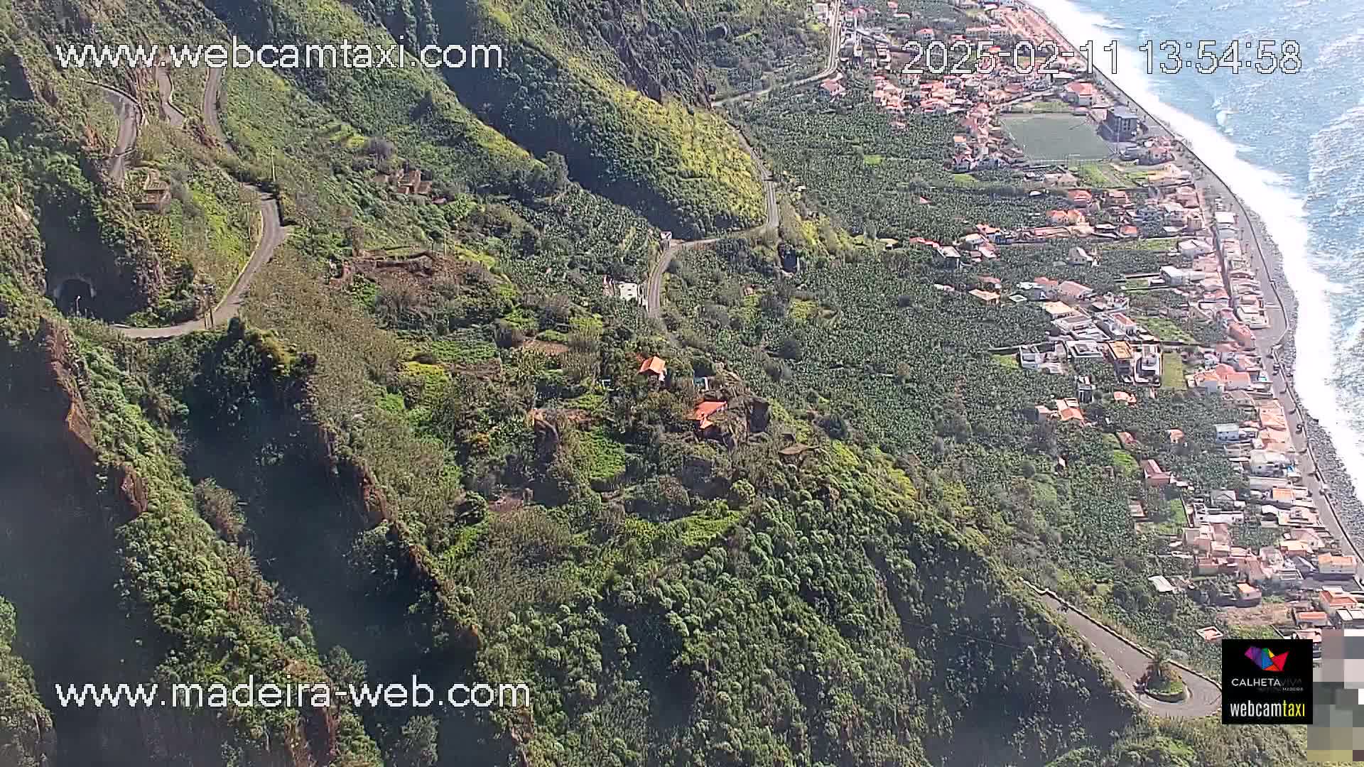 A high-angle view shows a winding road traversing a lush, green, mountainous landscape next to a coastline with a village and ocean under sunny conditions.